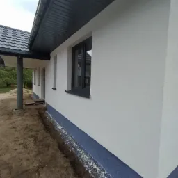 Świeżo pomalowana elewacja domu jednorodzinnego w kolorze białym, z ciemnoszarą podmurówką i stolarką okienną, widok z boku na tle zieleni.