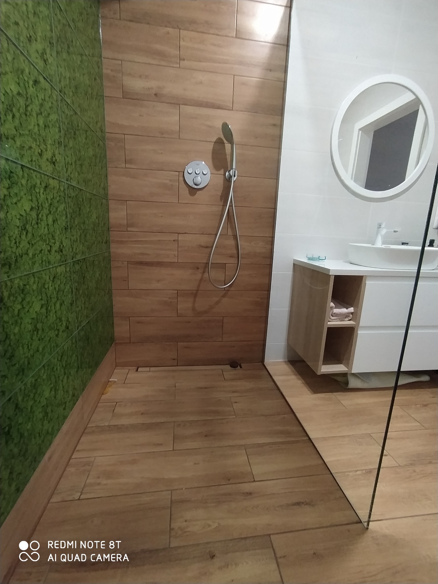 Nowoczesna kabina prysznicowa walk-in z zieloną ścianą z płytek imitujących mech i drewnopodobnymi płytkami na pozostałych ścianach, okrągłe lustro nad białą szafką z umywalką.