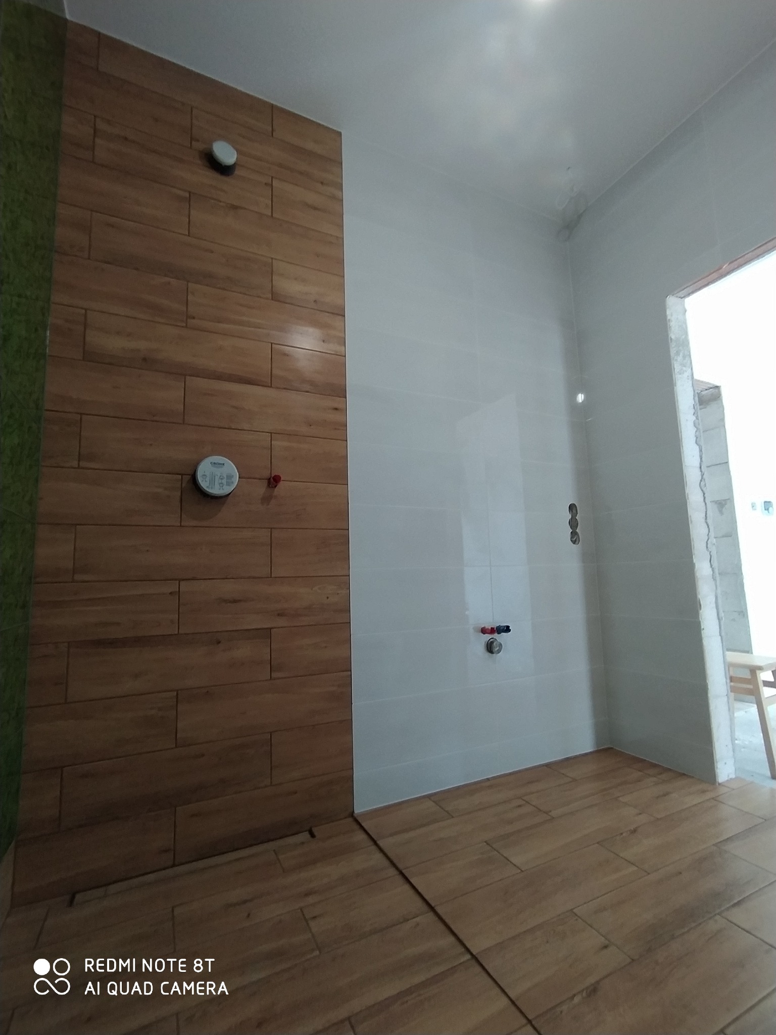 Nowoczesna łazienka w trakcie remontu: ściana z płytek imitujących drewno, białe płytki na pozostałych ścianach, widoczne instalacje wodne i otwór drzwiowy w stanie surowym.