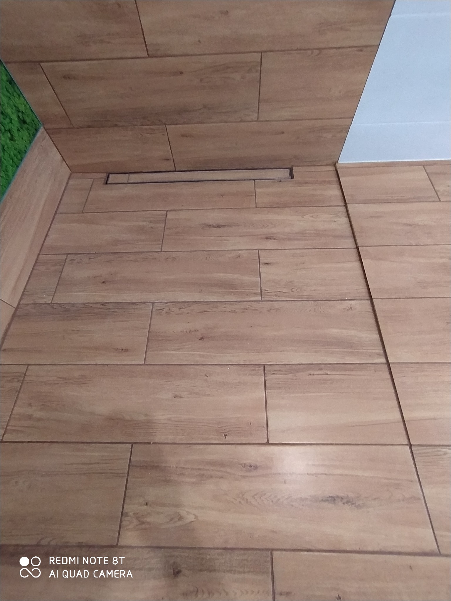 Ułożone płytki podłogowe imitujące drewno w łazience z odpływem liniowym, widoczny fragment ściany wykończonej na biało i sztucznej trawy.