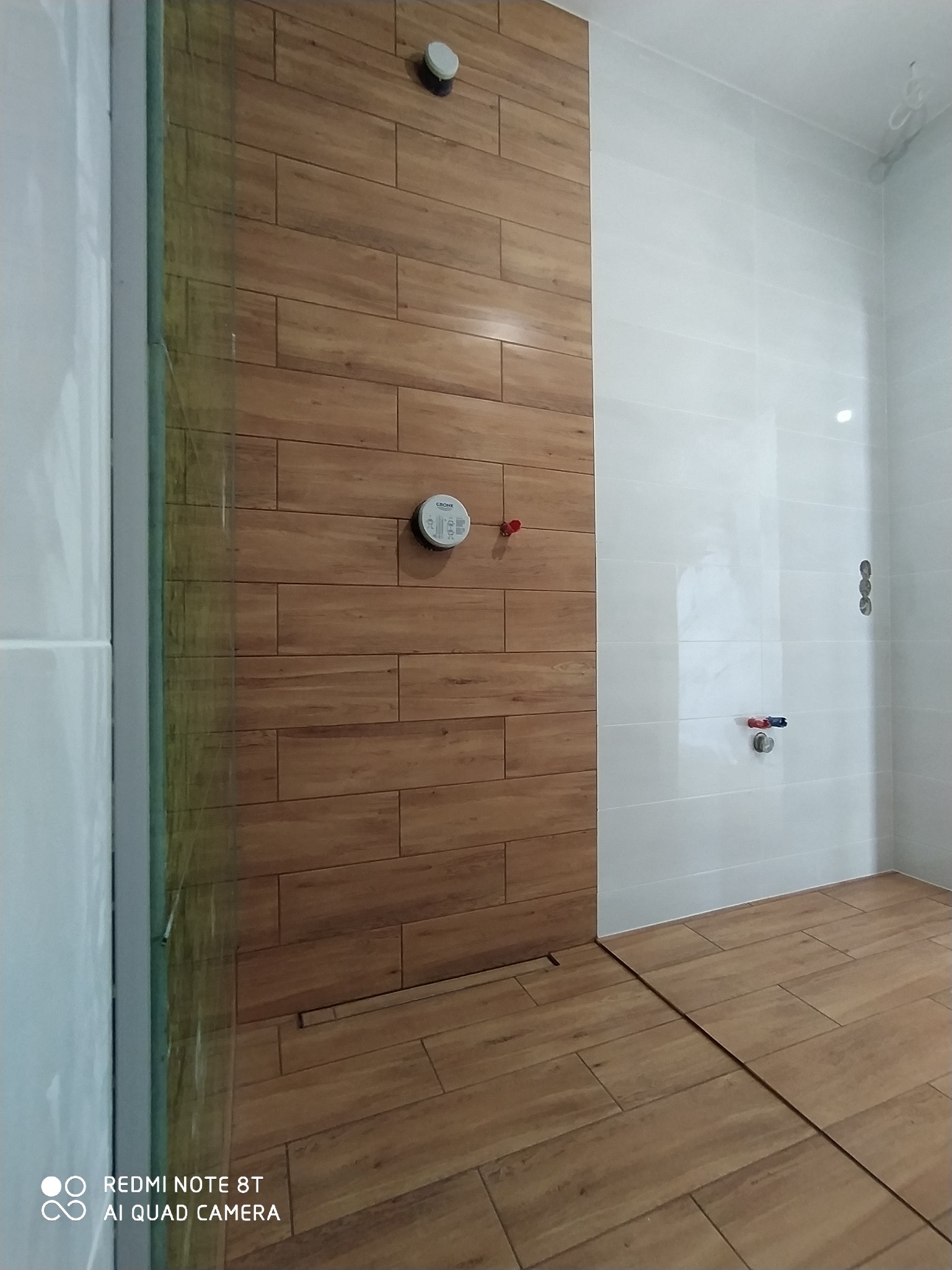 Nowoczesna kabina prysznicowa z drewnopodobnymi płytkami na ścianie i podłodze, szklana przegroda, białe płytki na pozostałych ścianach, widoczny odpływ liniowy.