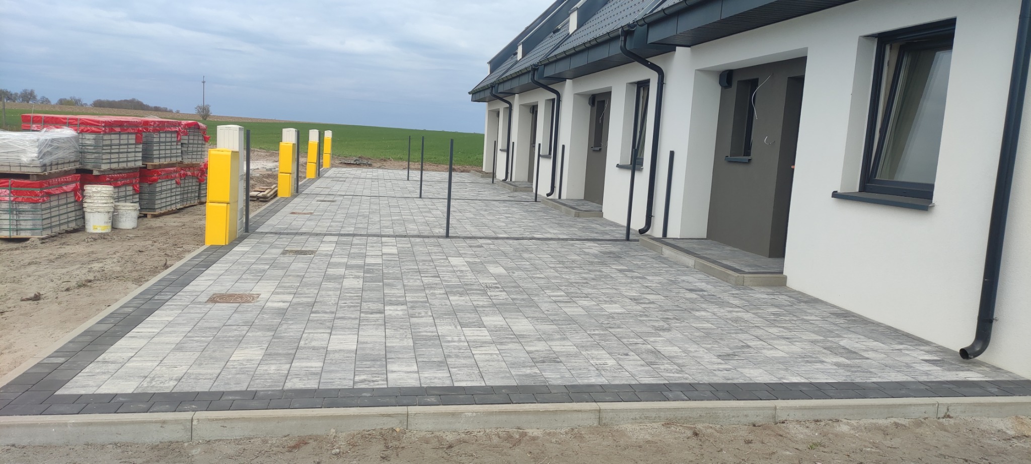 Ułożona szara kostka brukowa przed szeregiem nowych domów z białymi elewacjami i szarymi drzwiami, widoczne palety z kostką brukową i żółte słupki zabezpieczające.