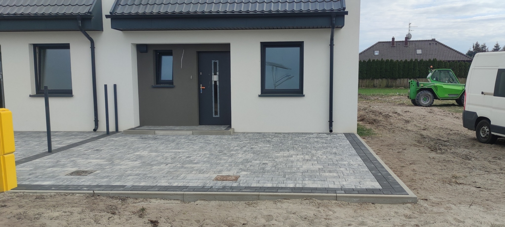 Nowoczesny dom bliźniak z szarą kostką brukową na podjeździe i chodniku, obrzeża z ciemnej kostki, w tle zielony wózek widłowy i częściowo widoczny biały bus