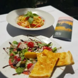 Dwa talerze: carpaccio z pomidorami i rukolą z cytryną oraz focaccia na pierwszym planie, spaghetti z krewetkami i bazylią w tle, obok menu z krajobrazem.