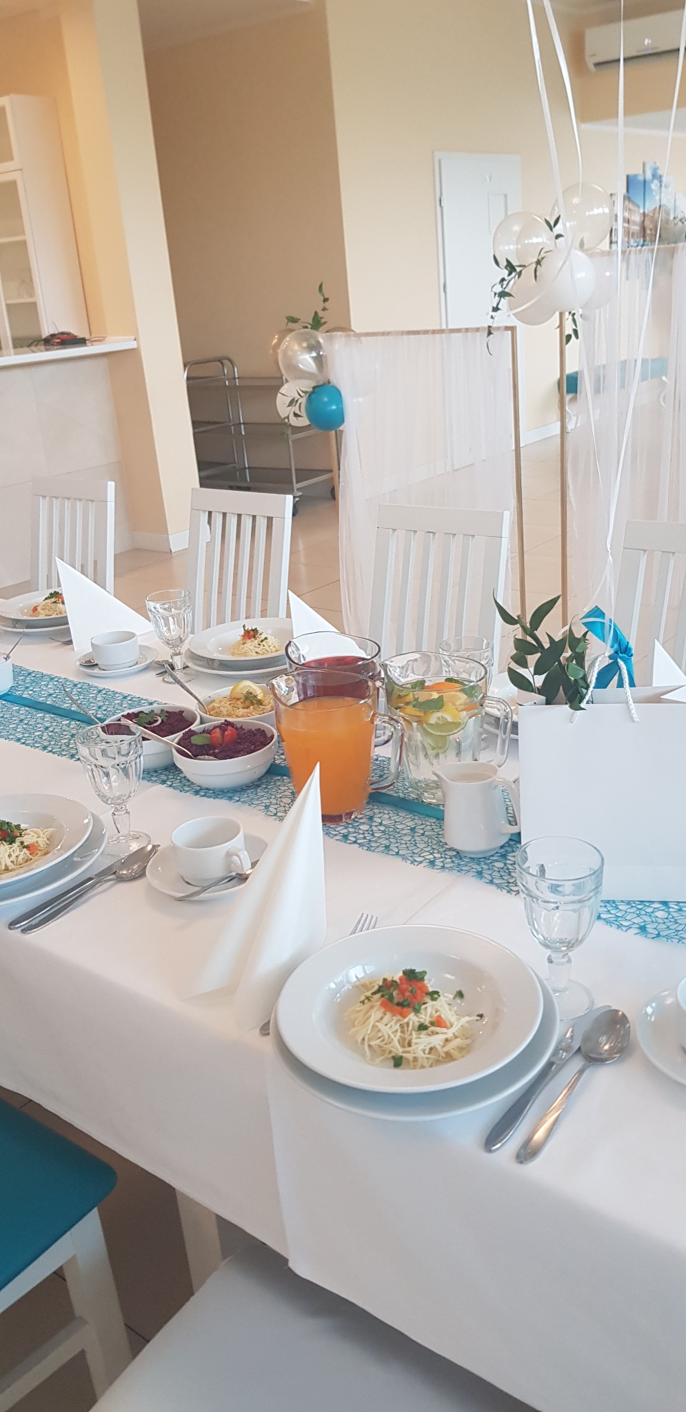 Uroczysty stół z białym obrusem, dekorowany błękitną bieżnikiem, zastawiony talerzami z daniami, szklankami, dzbankami z napojami, białymi serwetkami i balonami w tle.