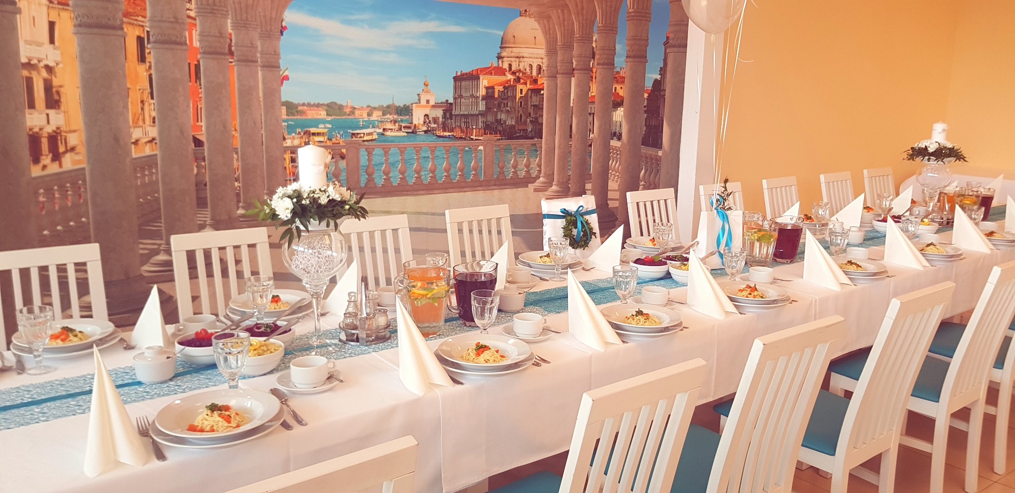 Długi stół z białym obrusem i turkusowym bieżnikiem, nakryty dla gości: talerze z makaronem, szklanki, napoje w dzbankach, białe serwetki ułożone w trójkąty, w tle fototapeta z weneckim krajobrazem...