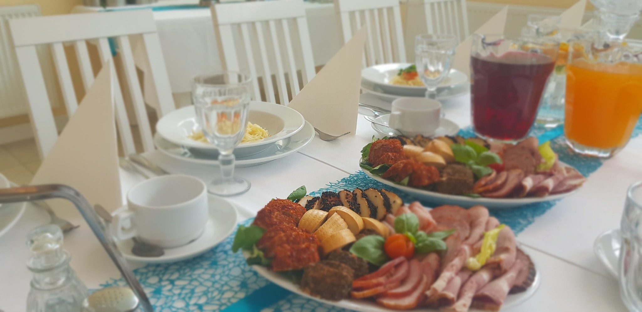 Uroczysty stół z dwoma talerzami wędlin i przekąsek, dzbankami z sokami, filiżankami, talerzami z makaronem i serwetkami, ustawiony na białym obrusie z turkusowym bieżnikiem.