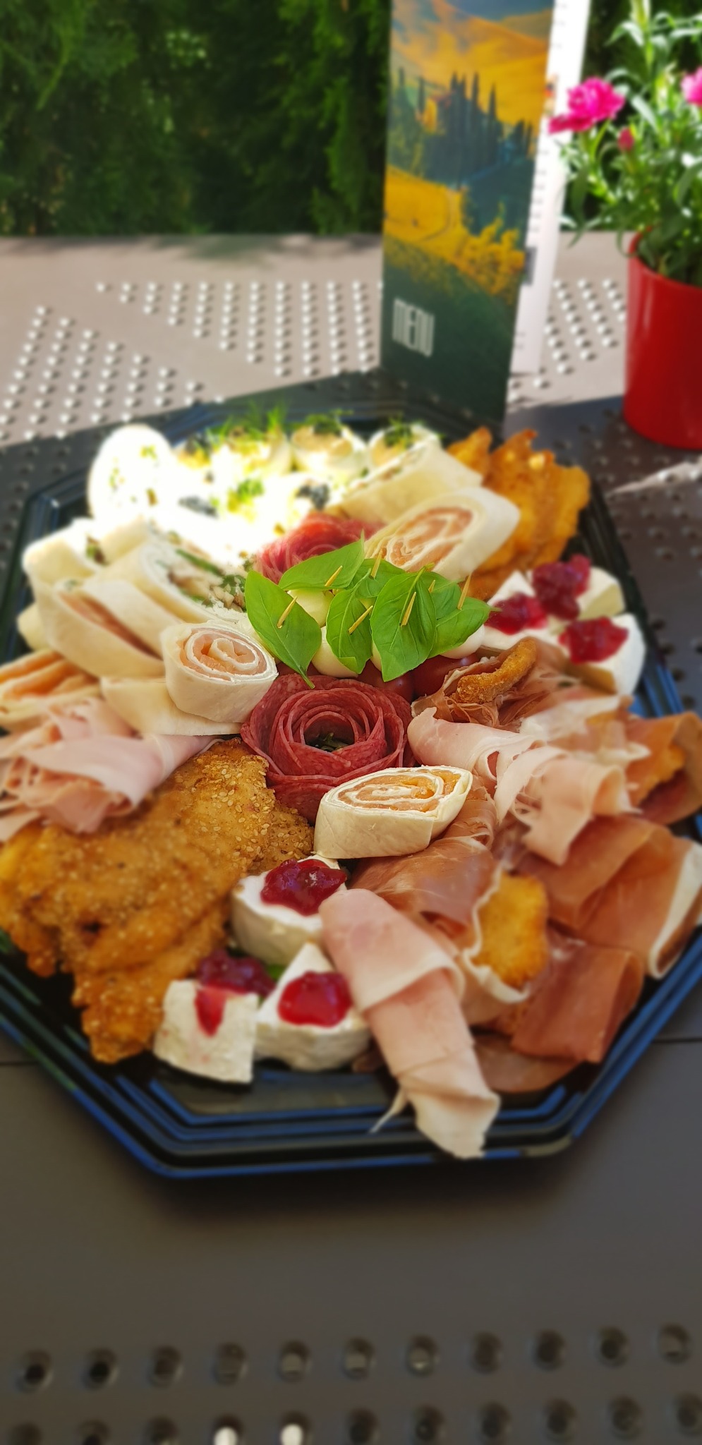 Elegancko ułożony catering na czarnym talerzu: rolady z szynki i sera, jajka faszerowane, ser z żurawiną, salami uformowane w róże, menu w tle i doniczka z kwiatami.