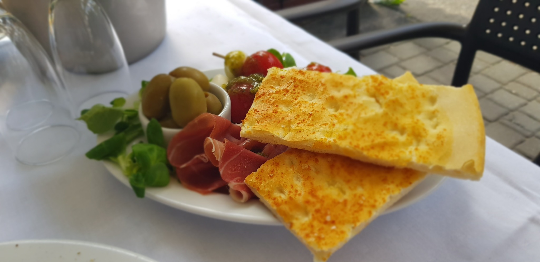 Przystawka z focaccią, szynką parmeńską, oliwkami i warzywami serwowana w Kluczborku na białym obrusie, z widocznymi kieliszkami w tle.