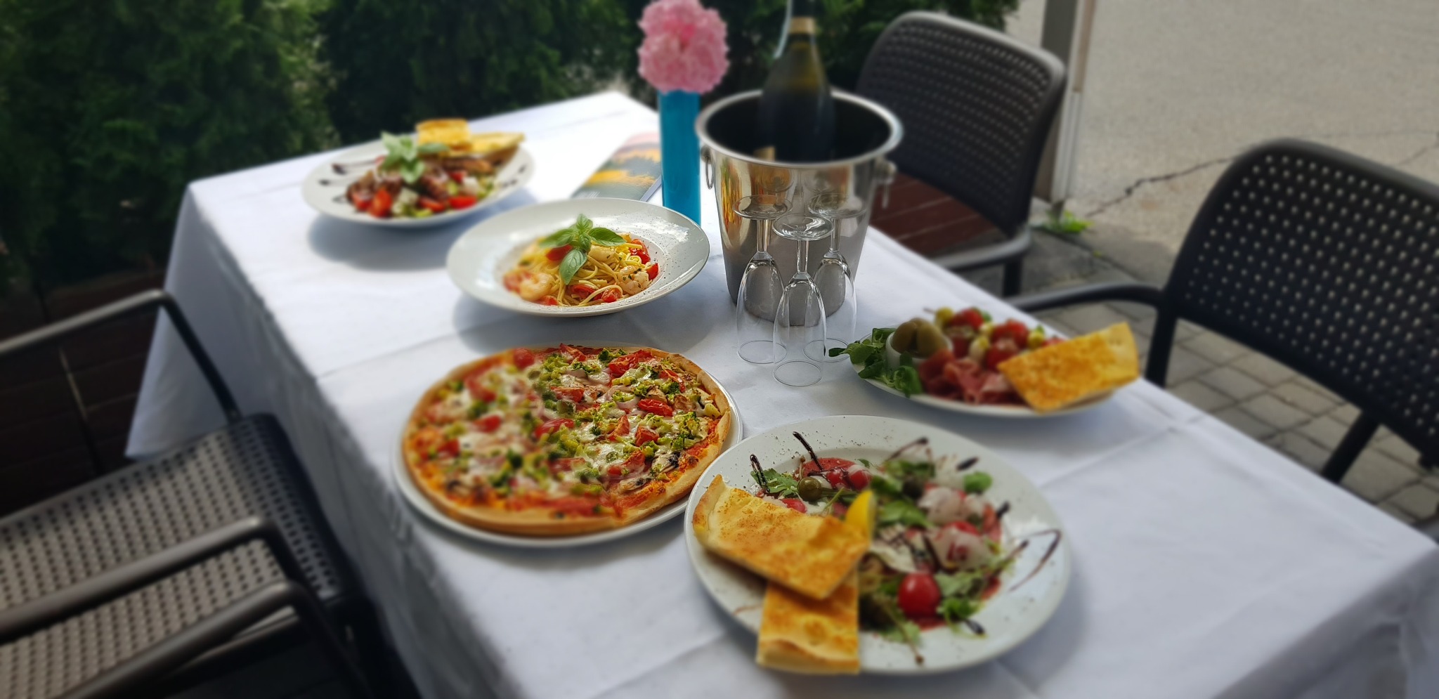 Stół nakryty białym obrusem z pizzą, spaghetti, sałatkami i przystawkami, butelka wina w wiaderku z lodem, ustawiony na zewnątrz restauracji.