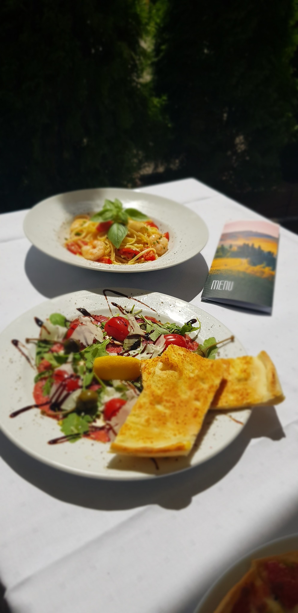 Dwa talerze: carpaccio z pomidorami i rukolą z cytryną oraz focaccia na pierwszym planie, spaghetti z krewetkami i bazylią w tle, obok menu z krajobrazem.