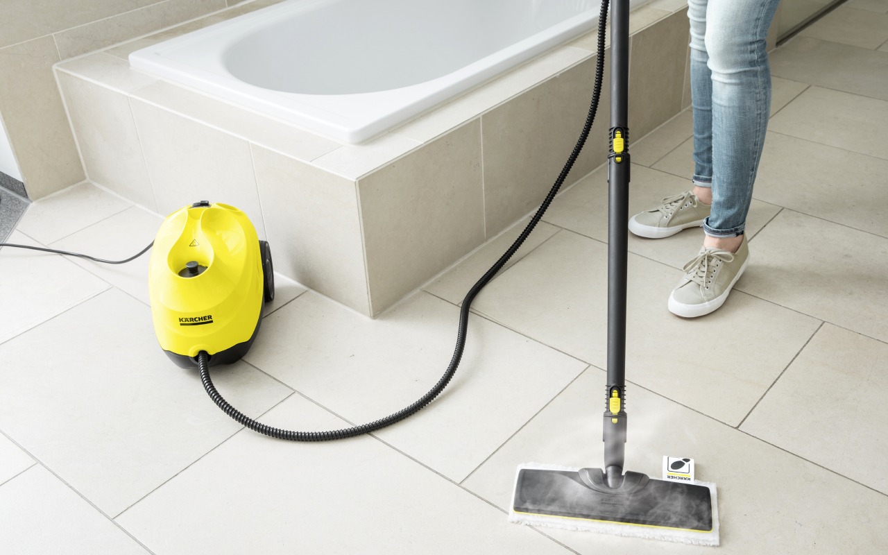 Żółty parowy mop Karcher w trakcie czyszczenia jasnych płytek podłogowych w łazience, widoczna para i fragment wanny.