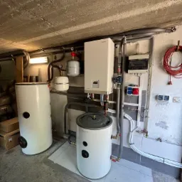 Dwa białe zbiorniki akumulacyjne ciepła z szarymi elementami, podłączone do instalacji grzewczej w pomieszczeniu z betonowym sufitem i szarą podłogą z płytek.