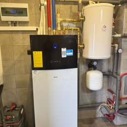 ECO CONNECT SPÓŁKA Z OGRANICZONĄ ODPOWIEDZIALNOŚCIĄ - Zainstalowana pompa ciepła Daikin w kotłowni z widocznymi rurami instalacji CO i CWU oraz zbiornikiem buforowym. Na ścianie skrzynka elektryczna i naklejki informacyjne.