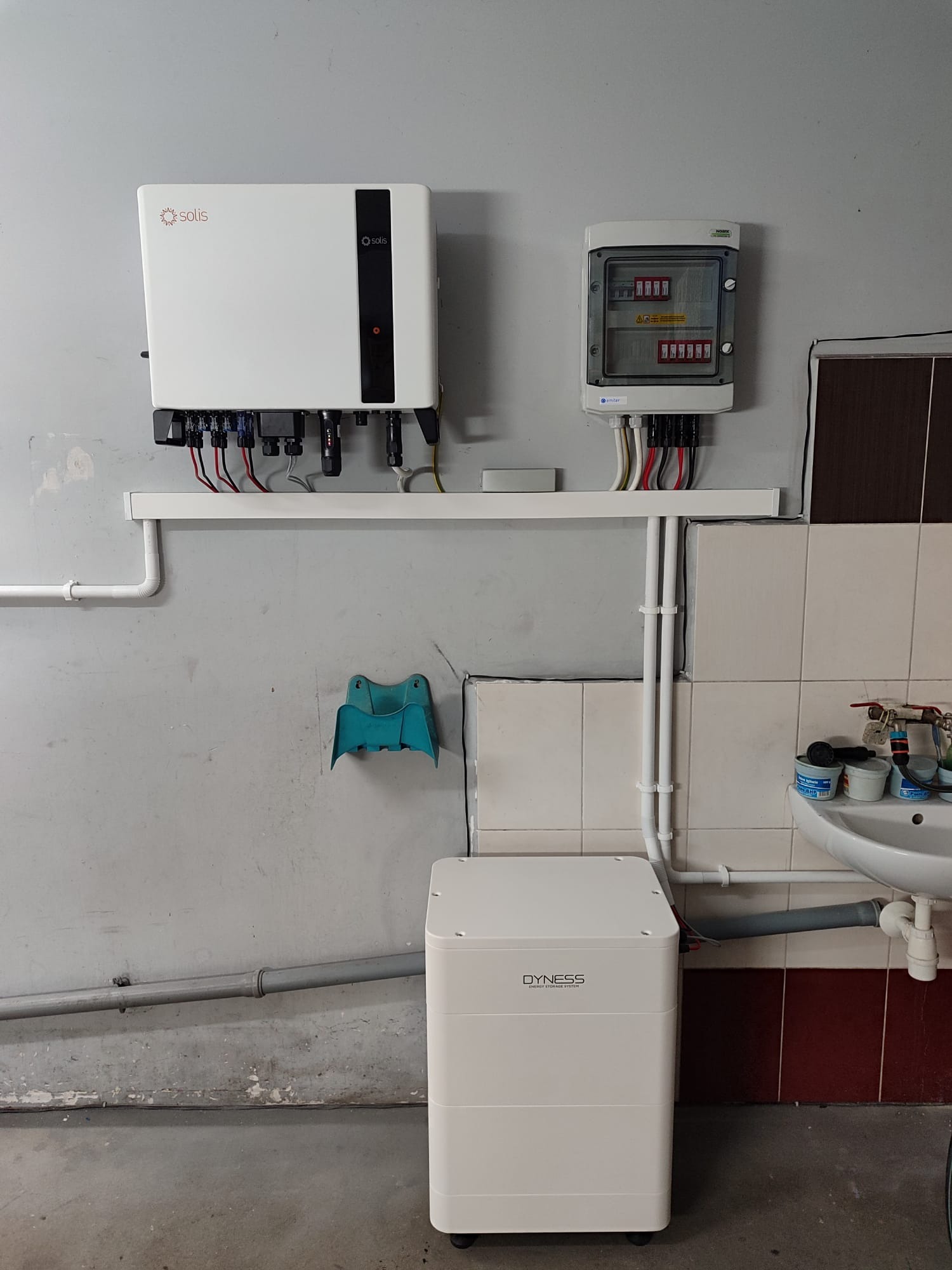 Instalacja fotowoltaiczna w pomieszczeniu: biały falownik Solis, rozdzielnica elektryczna w obudowie i akumulator Dyness, zamontowane na ścianie z widocznymi rurami i umywalką.