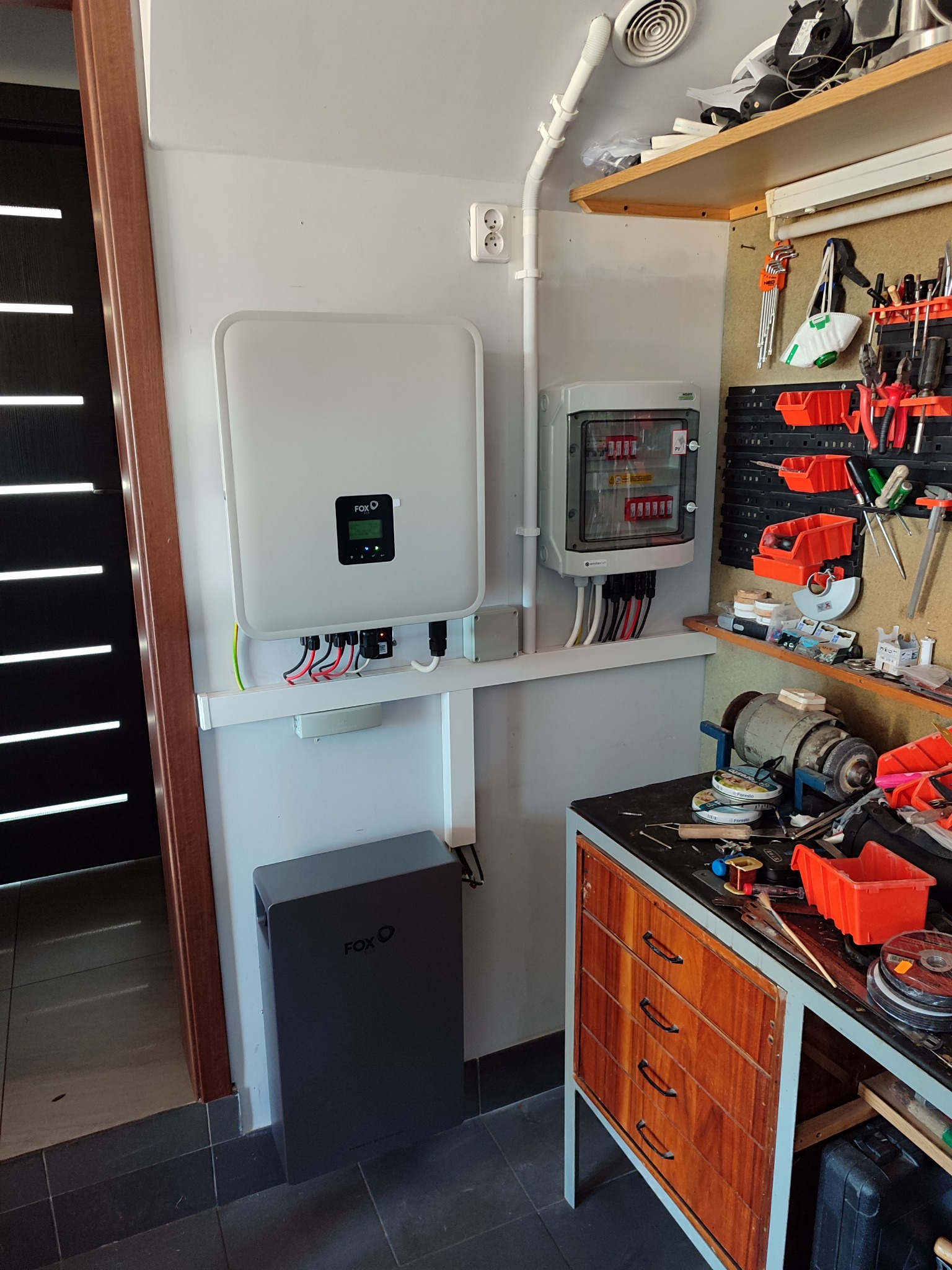 Wnętrze pomieszczenia z zamontowanym inwerterem i akumulatorem energii firmy FoxESS, skrzynką zabezpieczeń oraz warsztatem z narzędziami w tle.
