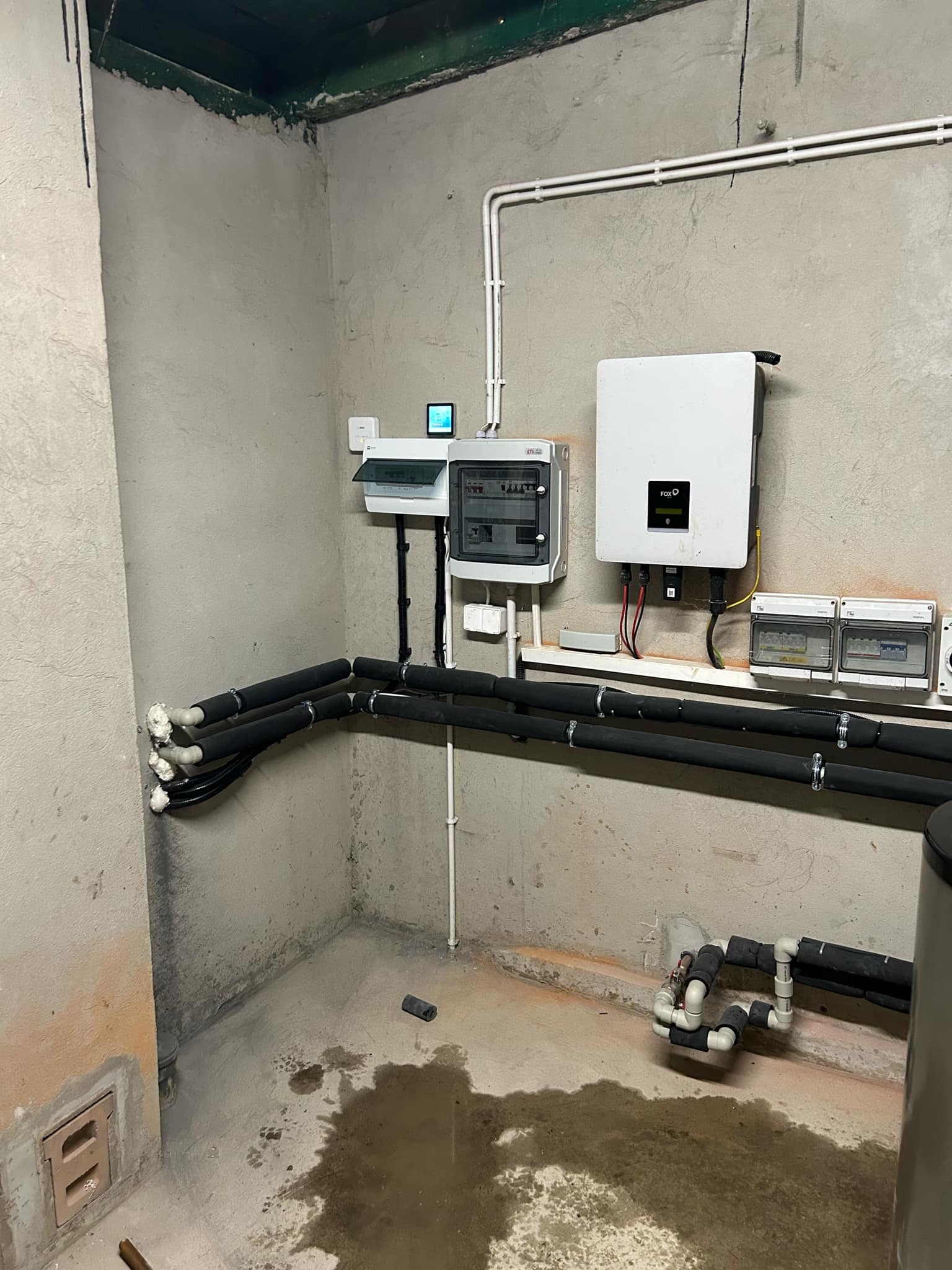 Instalacja z czarnymi, izolowanymi rurami, białą skrzynką rozdzielczą i falownikiem FOX ESS na szarej, surowej ścianie w pomieszczeniu technicznym z wilgotną podłogą.