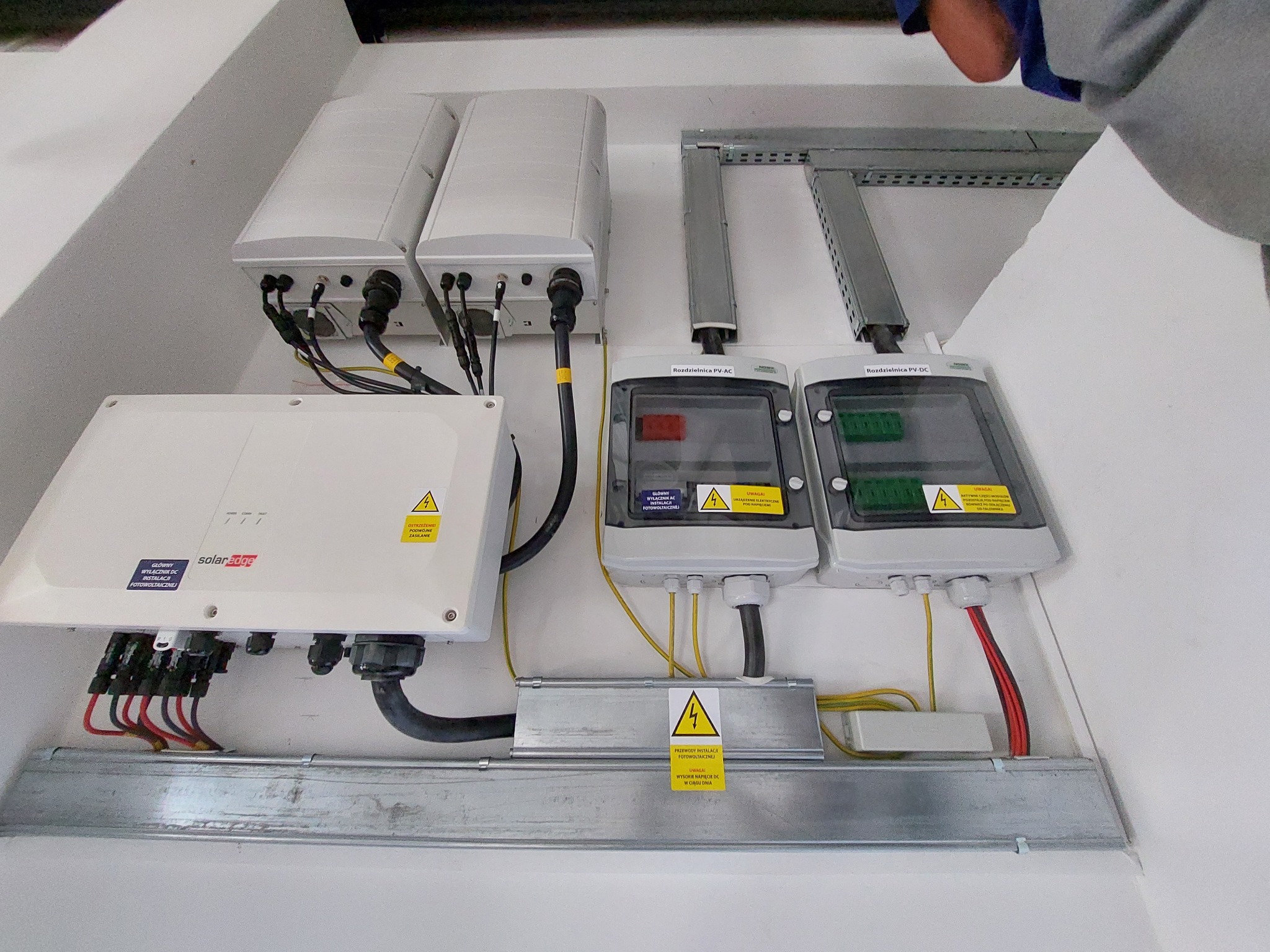 Szczegółowe ujęcie instalacji fotowoltaicznej wewnątrz budynku, widoczne moduły falownika SolarEdge, rozdzielnice PV-AC i PV-DC, okablowanie w korytkach oraz ostrzeżenia o napięciu.