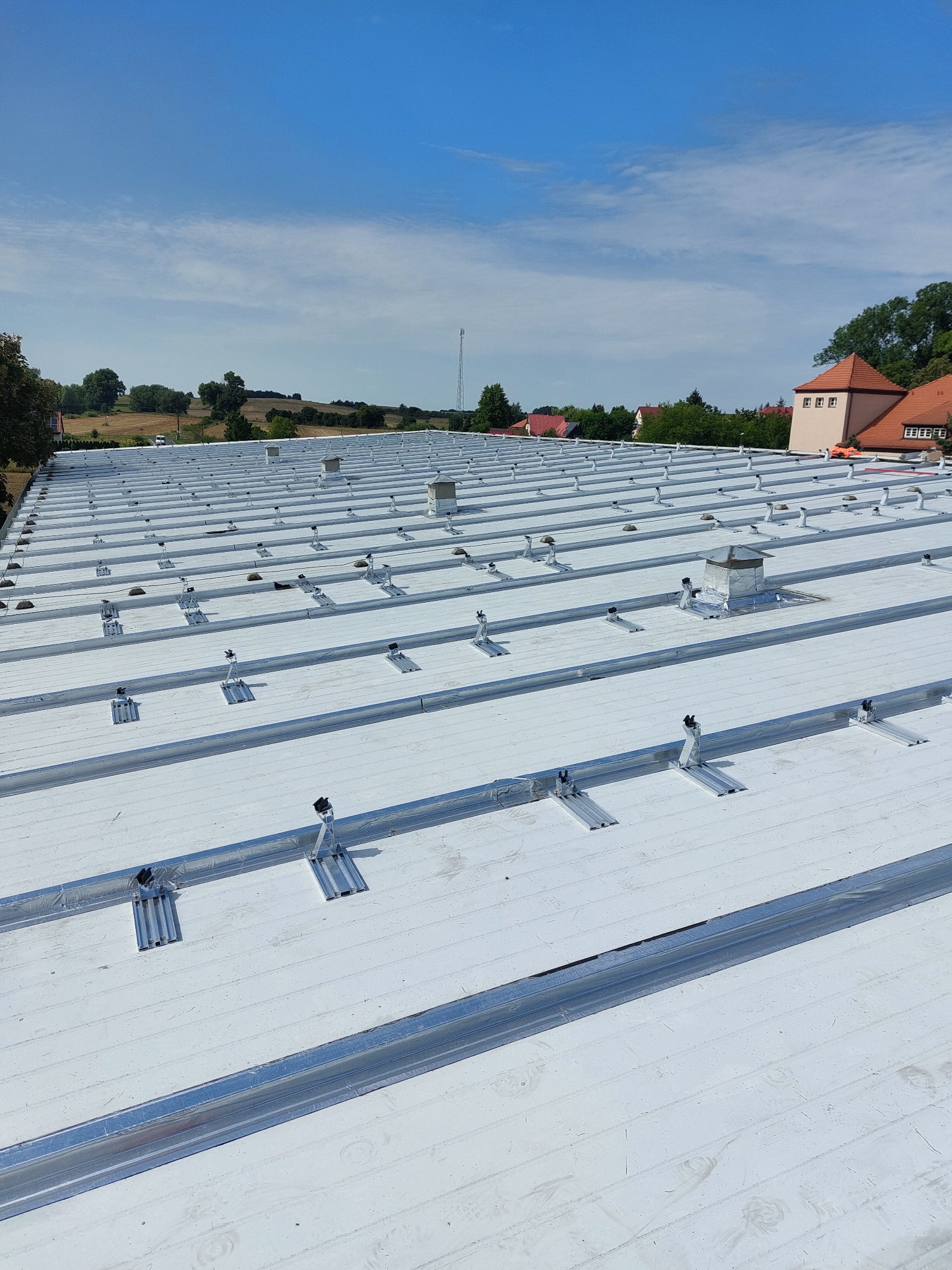 Biały dach z zamontowanymi szynami montażowymi pod panele fotowoltaiczne, widok z góry na tle błękitnego nieba i zabudowań.