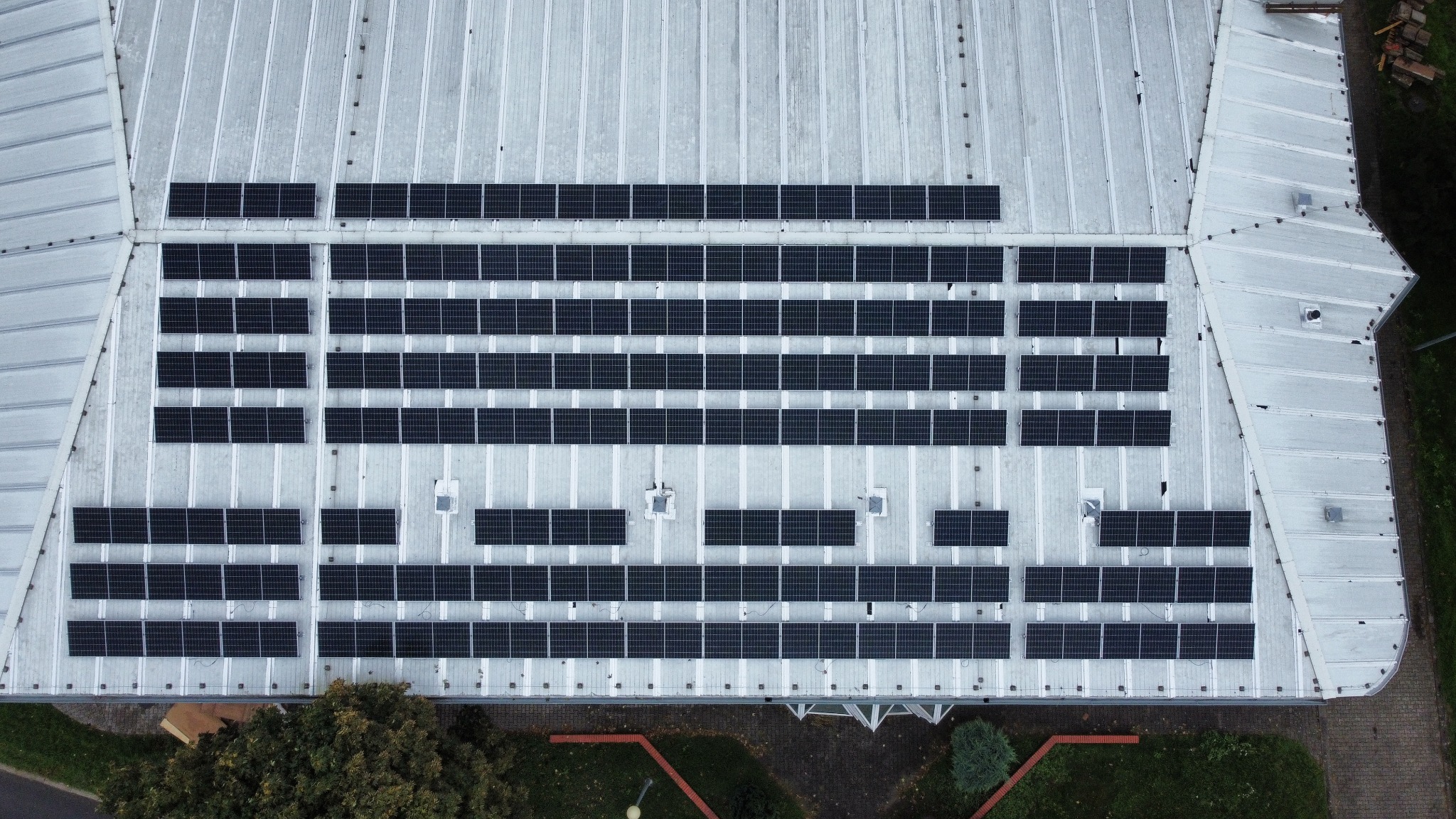 Widok z drona na duży dach pokryty panelami fotowoltaicznymi, ułożonymi w rzędach na szarym, metalowym dachu budynku przemysłowego.
