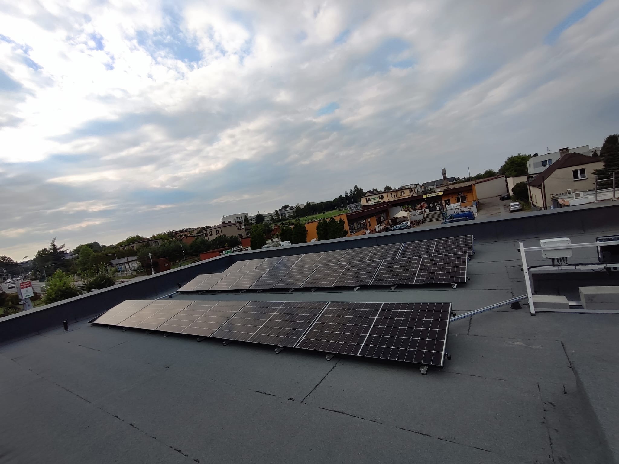 Dwa rzędy paneli fotowoltaicznych na płaskim dachu budynku w pochmurny dzień, widok z góry na tle zabudowy miejskiej.