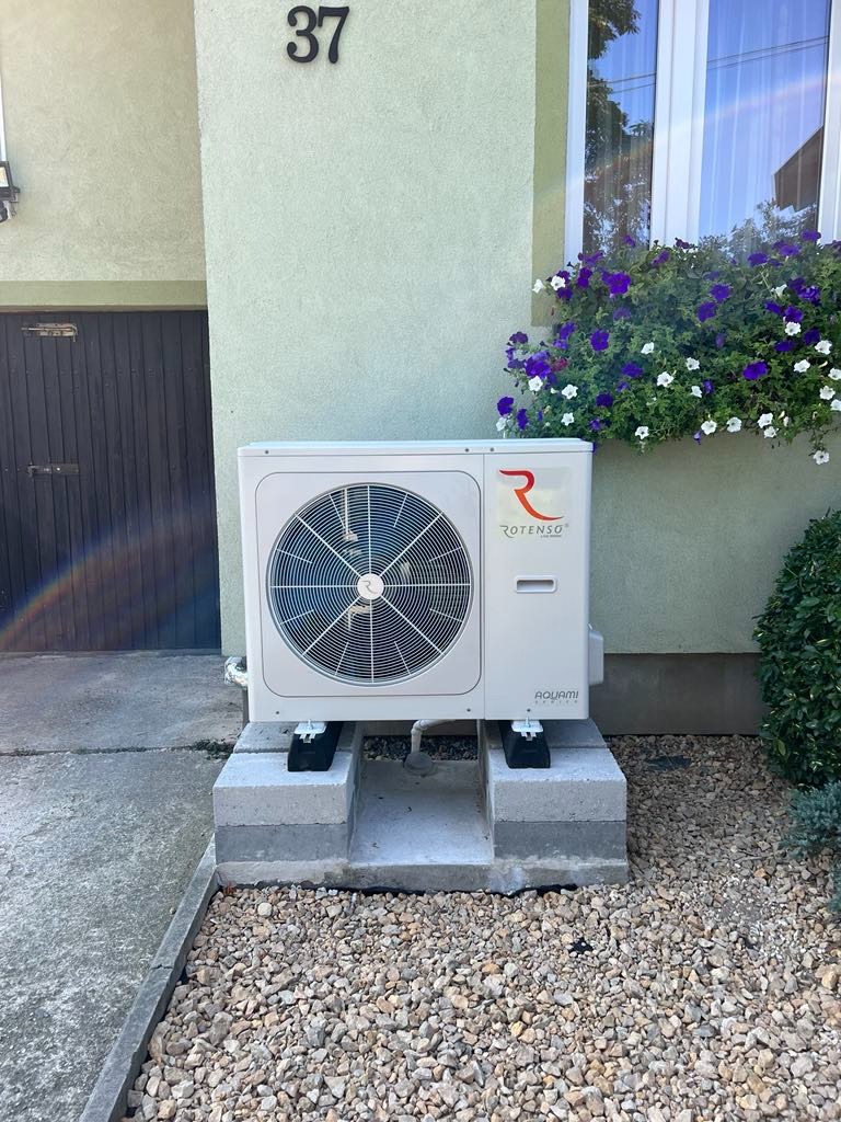 Biała pompa ciepła Rotenso Aquami stojąca na betonowych bloczkach przed budynkiem z numerem 37, z kwiatami w oknie powyżej.