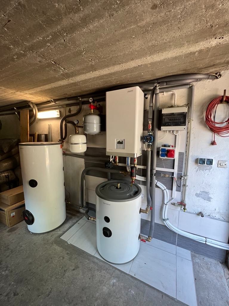 Dwa białe zbiorniki akumulacyjne ciepła z szarymi elementami, podłączone do instalacji grzewczej w pomieszczeniu z betonowym sufitem i szarą podłogą z płytek.