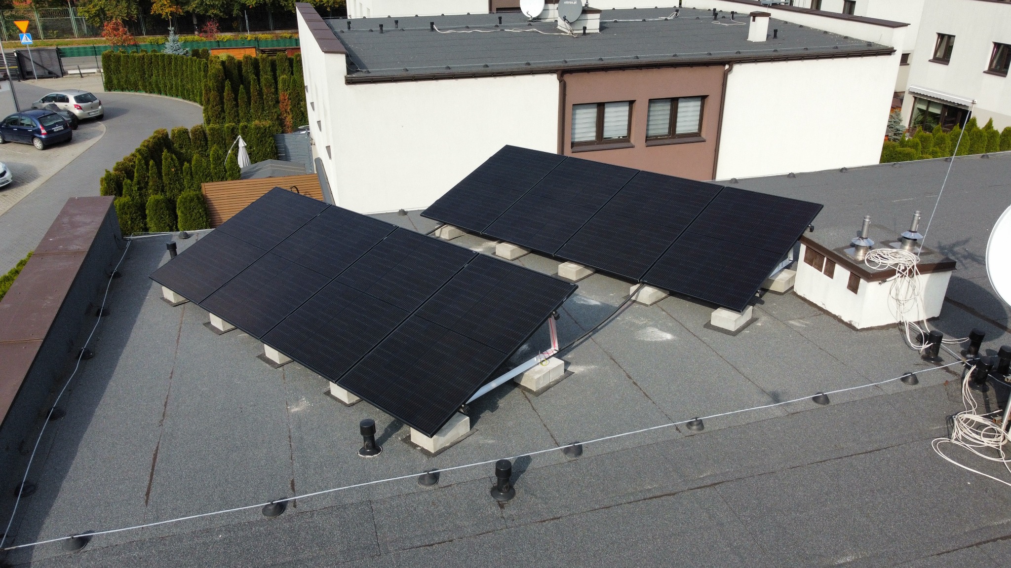 Dwa rzędy paneli słonecznych na płaskim dachu budynku mieszkalnego, widok z góry, w tle zabudowa osiedlowa i zieleń.