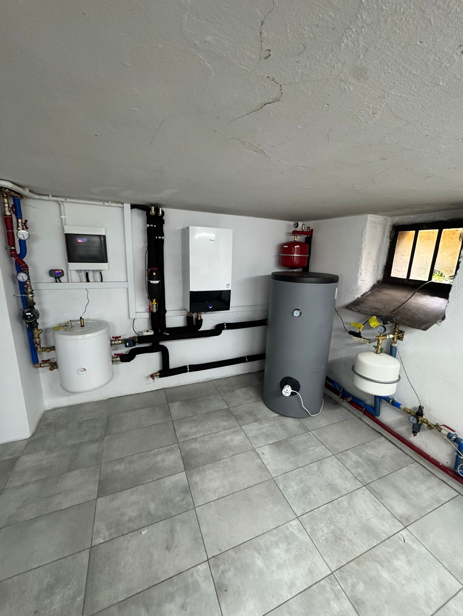Zestaw urządzeń grzewczych w kotłowni: zbiornik akumulacyjny, bufor ciepła, pompa obiegowa i rozdzielnica. Instalacja z rur miedzianych i czarnych. Wykończona posadzka.