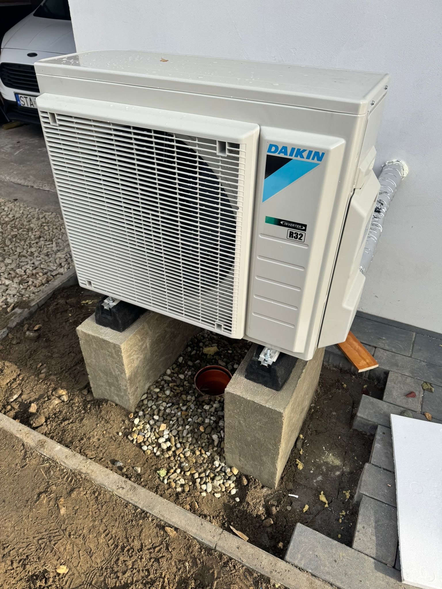 Zewnętrzna jednostka pompy ciepła Daikin, model Inverter R32, zamontowana na betonowych blokach w przygotowanym wykopie. Widoczne elementy instalacji i otoczenie.