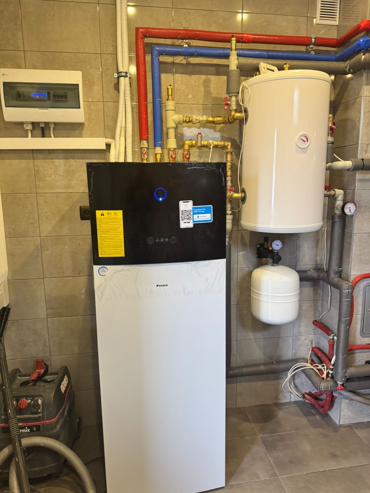 Zainstalowana pompa ciepła Daikin w kotłowni z widocznymi rurami instalacji CO i CWU oraz zbiornikiem buforowym. Na ścianie skrzynka elektryczna i naklejki informacyjne.