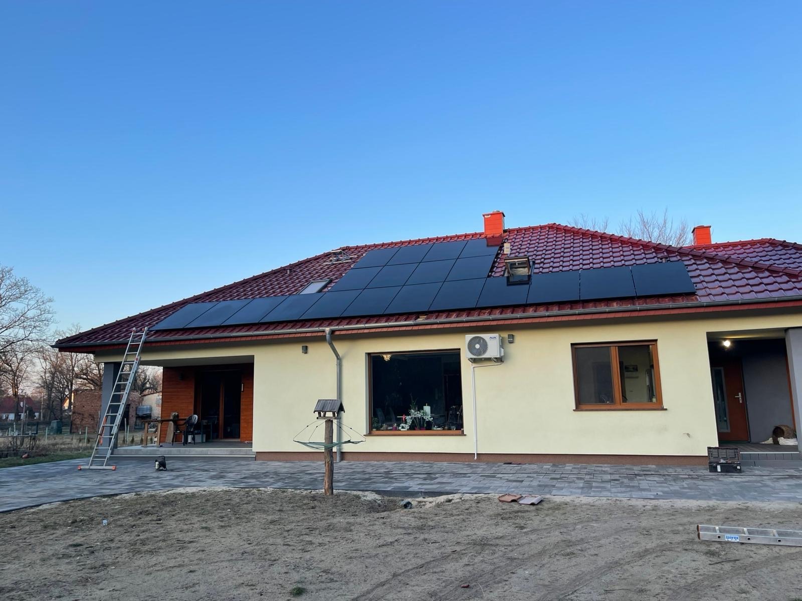 Dom jednorodzinny z panelami fotowoltaicznymi na dachu z czerwonej dachówki. Na ścianie zewnętrzna jednostka klimatyzacji. Drabina oparta o ścianę. Błękitne niebo.