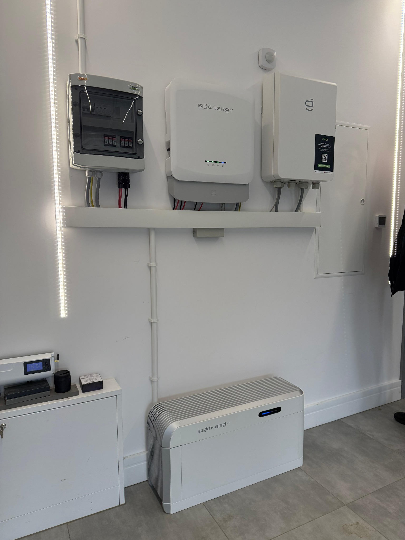 Biała instalacja z rozdzielnicą, inwerterem i akumulatorem na ścianie, z widocznymi przewodami i elementami montażowymi. Nowoczesny system zasilania.