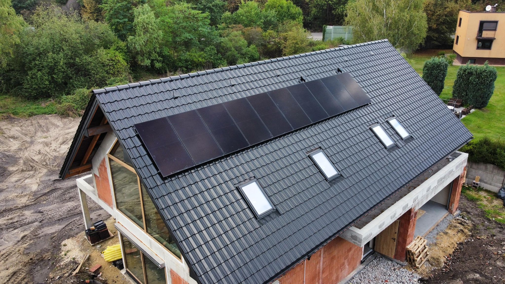 Czarny dach z panelami fotowoltaicznymi i oknami dachowymi na domu w trakcie budowy, widok z góry. W tle zieleń drzew.