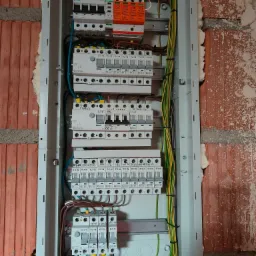 Rozdzielnica elektryczna z zamontowanymi zabezpieczeniami, widoczne przewody i aparatura, zamontowana na ścianie z czerwonej cegły. Widok z bliska.