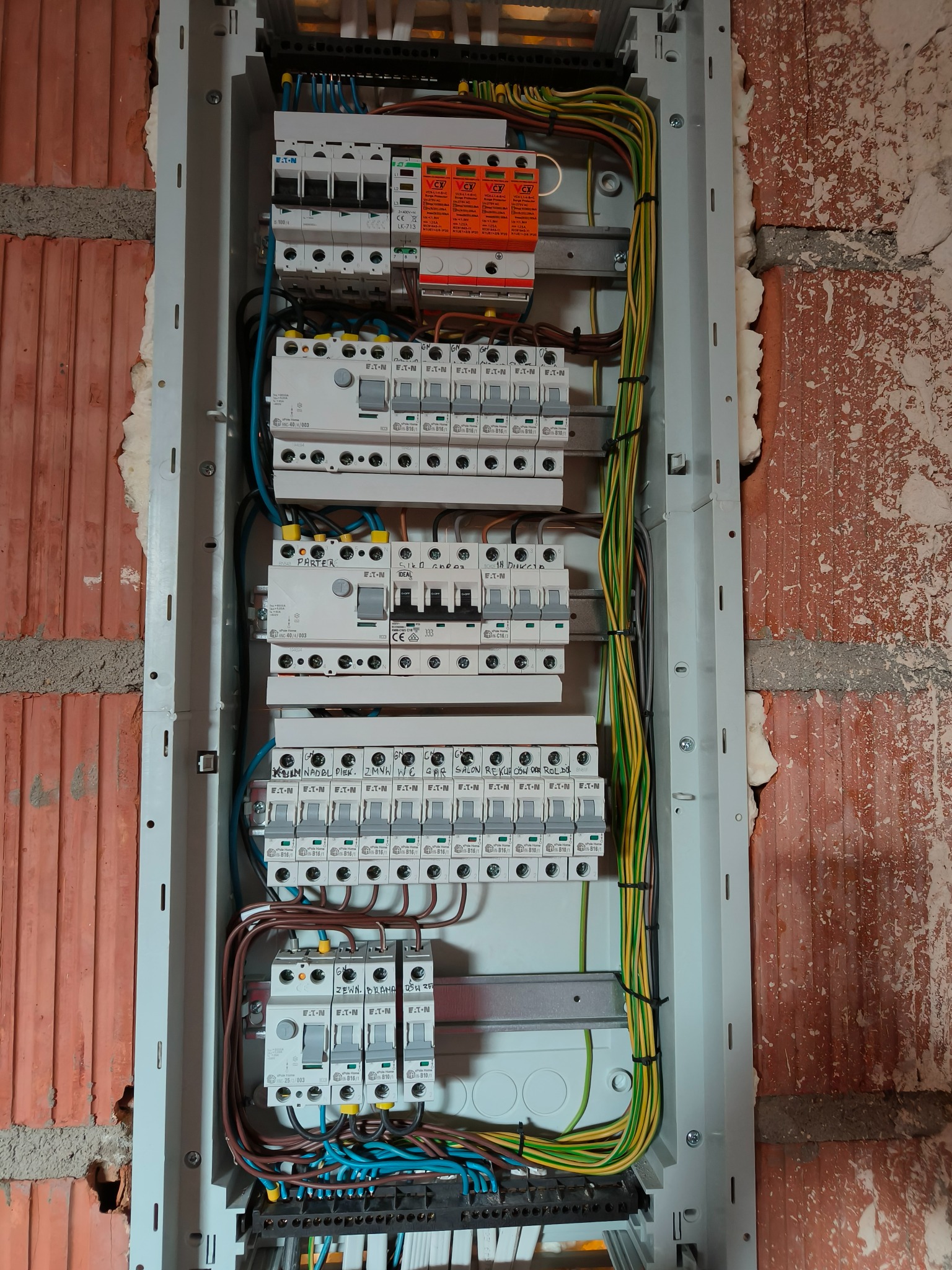 Rozdzielnica elektryczna z zamontowanymi zabezpieczeniami, widoczne przewody i aparatura, zamontowana na ścianie z czerwonej cegły. Widok z bliska.