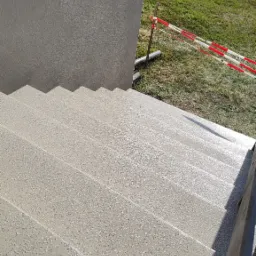Schody zewnętrzne pokryte jasną, szarą, chropowatą powłoką, zabezpieczone czerwoną taśmą ostrzegawczą, widok z góry.