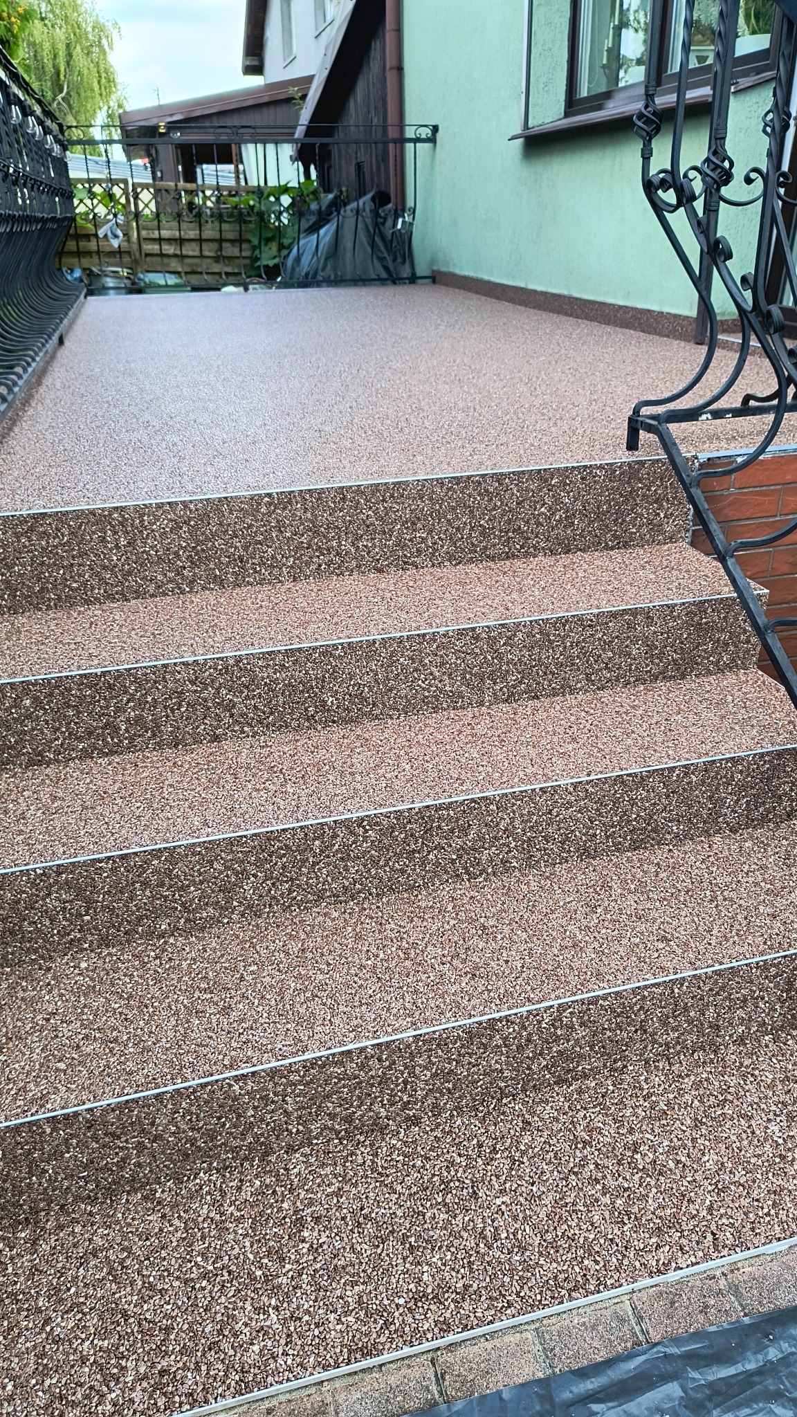 Schody i taras wykończone drobnym, brązowo-beżowym kruszywem z metalowymi listwami krawędziowymi, widok z dołu na górę.