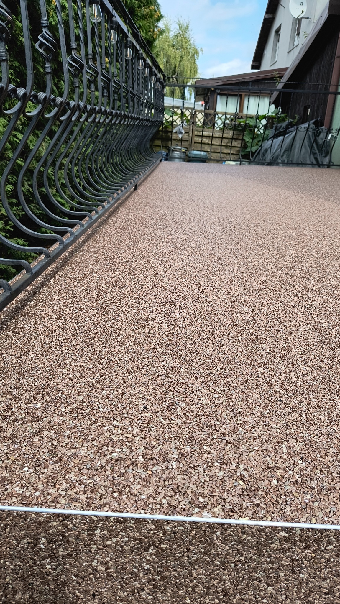 Beżowa, drobnoziarnista nawierzchnia balkonu z kutą, czarną balustradą, widok z góry, na zewnątrz.