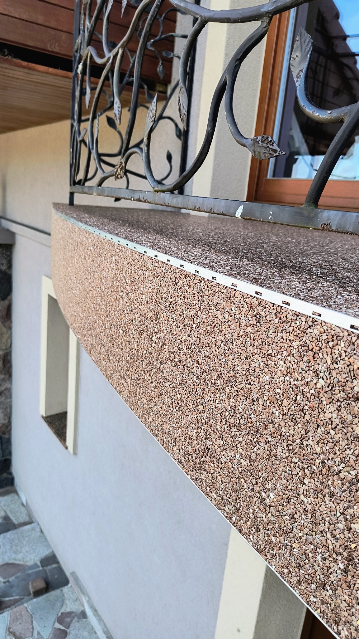 Wykończony balkon z posadzką żywiczną w odcieniach brązu, z ozdobną, metalową balustradą i widocznym fragmentem elewacji budynku.