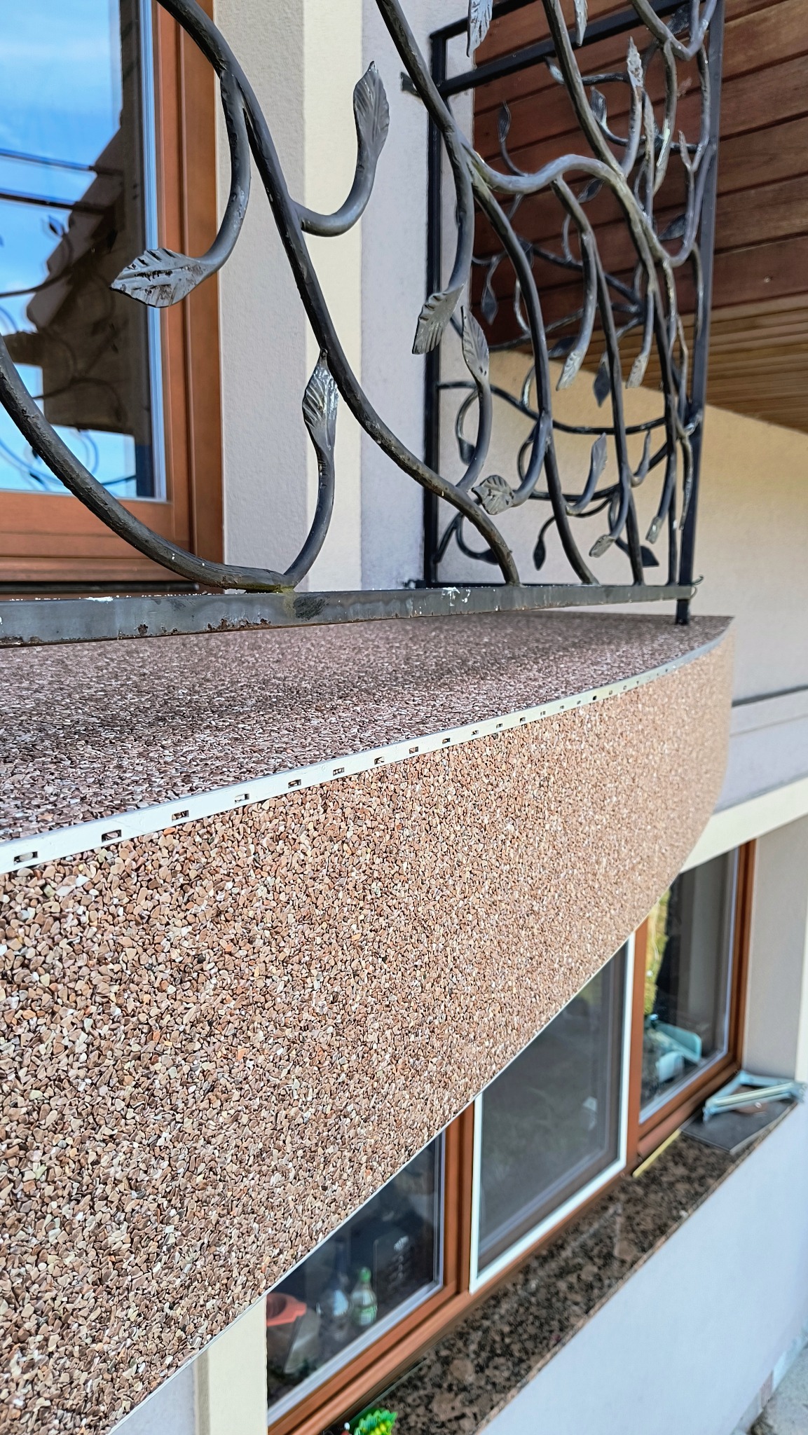 Wykończony balkon z brązową, kamyczkową powierzchnią, ozdobną metalową balustradą i drewnianymi ramami okiennymi.