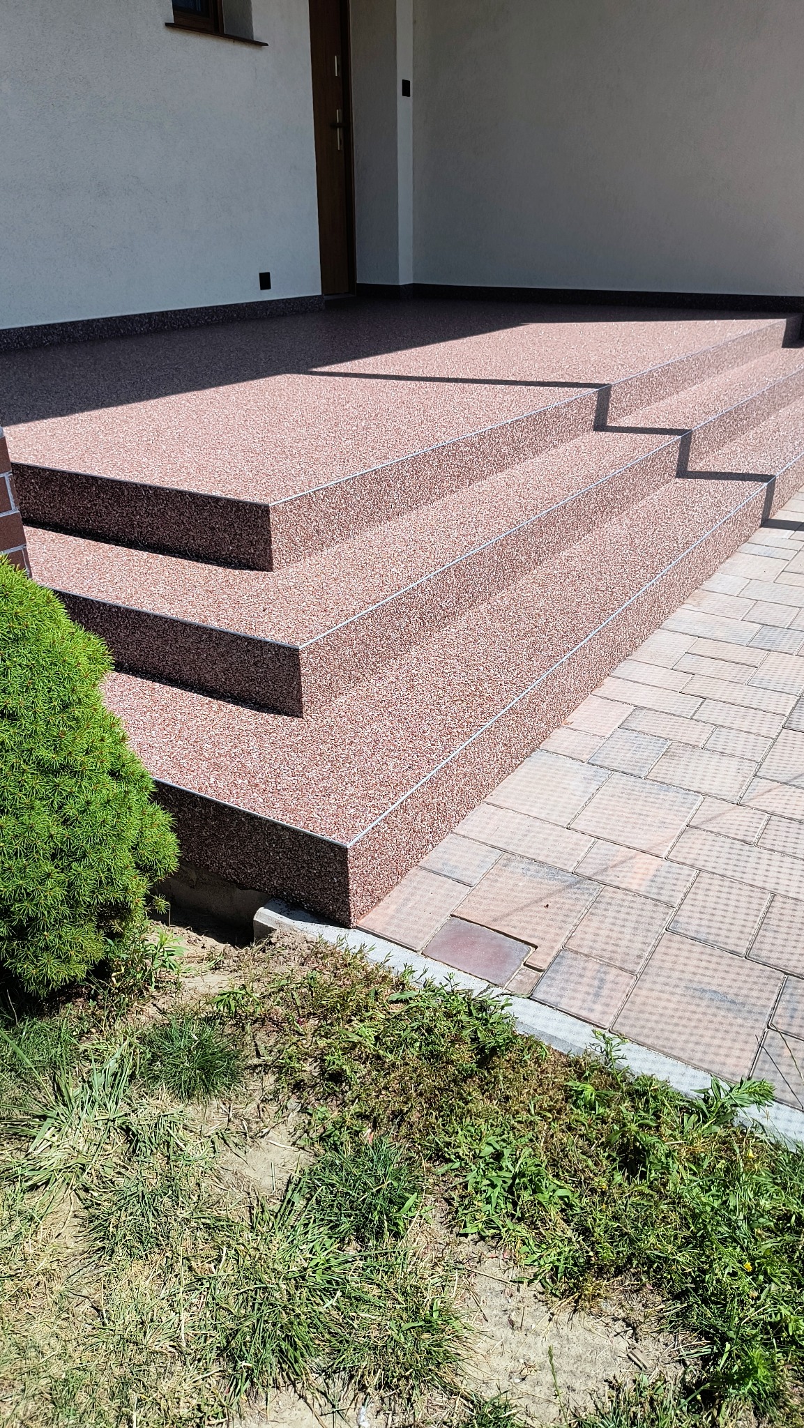 Zewnętrzne schody wejściowe pokryte brązowo-beżową, kamyczkową nawierzchnią, wykończone srebrnymi listwami, z widocznym fragmentem trawnika i kostki brukowej.