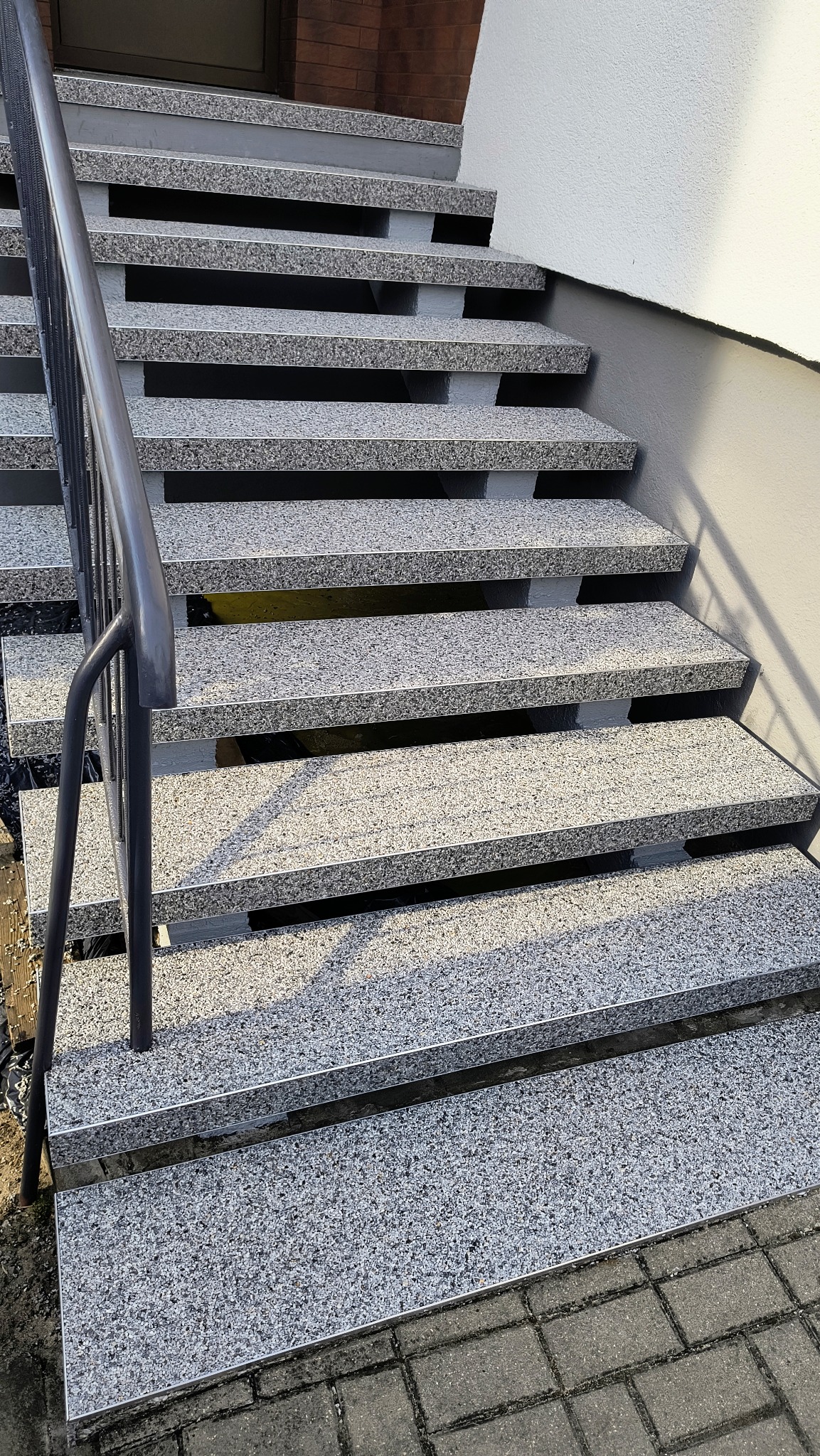 Zewnętrzne schody wykończone szarą, drobnoziarnistą posadzką z żywicy, z metalową barierką po lewej stronie i fragmentem chodnika z kostki brukowej u dołu.