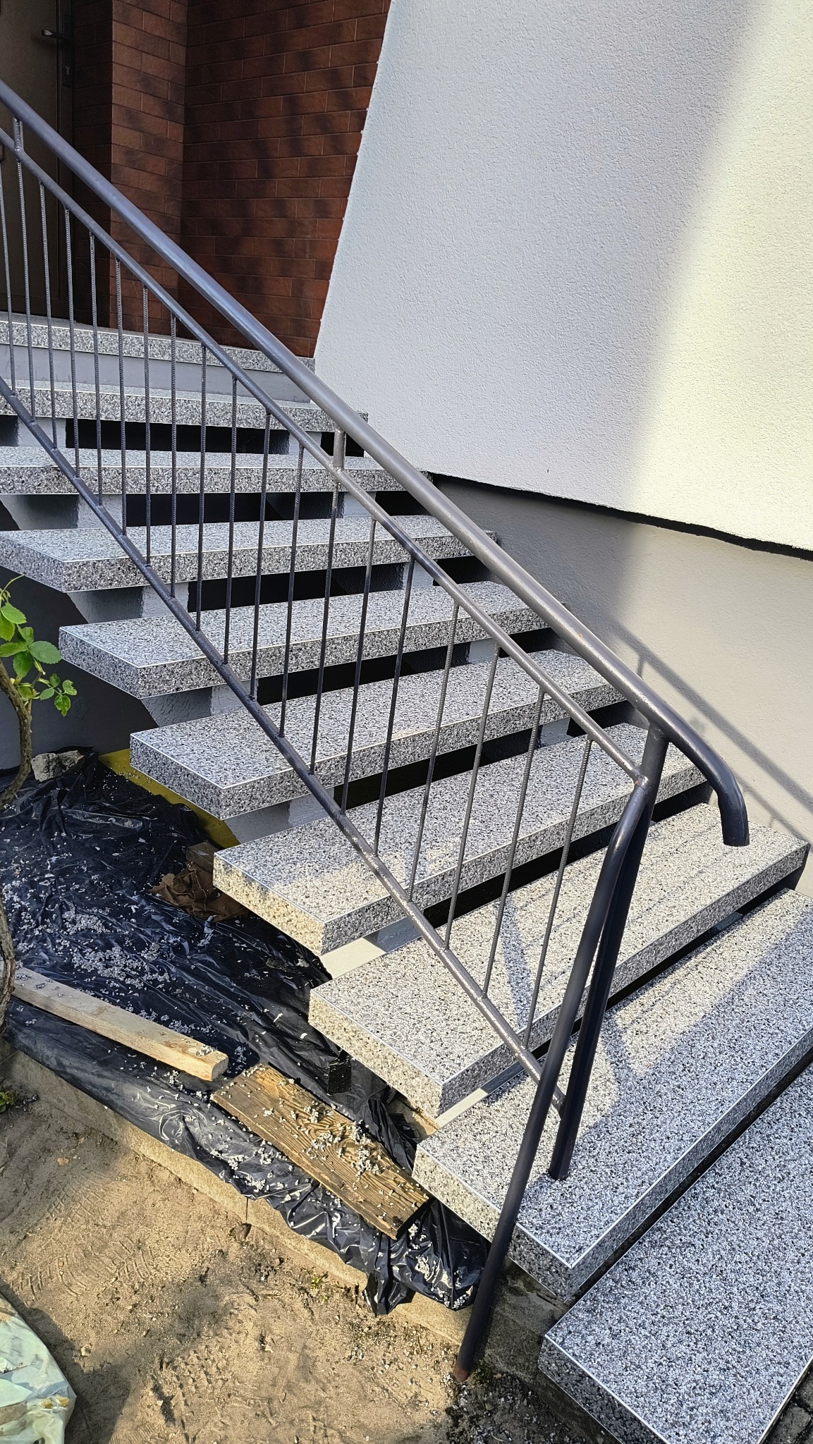 Zewnętrzne schody z szarego kamienia z metalową balustradą, widoczne ślady prac wykończeniowych i resztki materiałów budowlanych.
