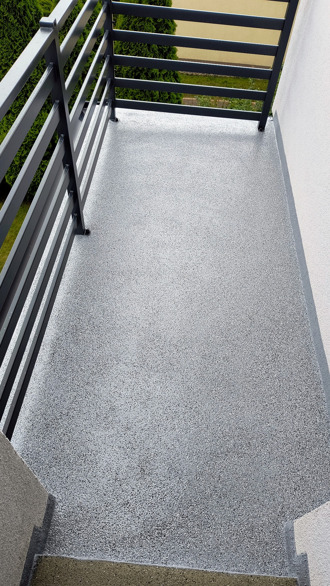 Szara, drobnoziarnista powierzchnia balkonu z metalową balustradą, widok z góry na schody.