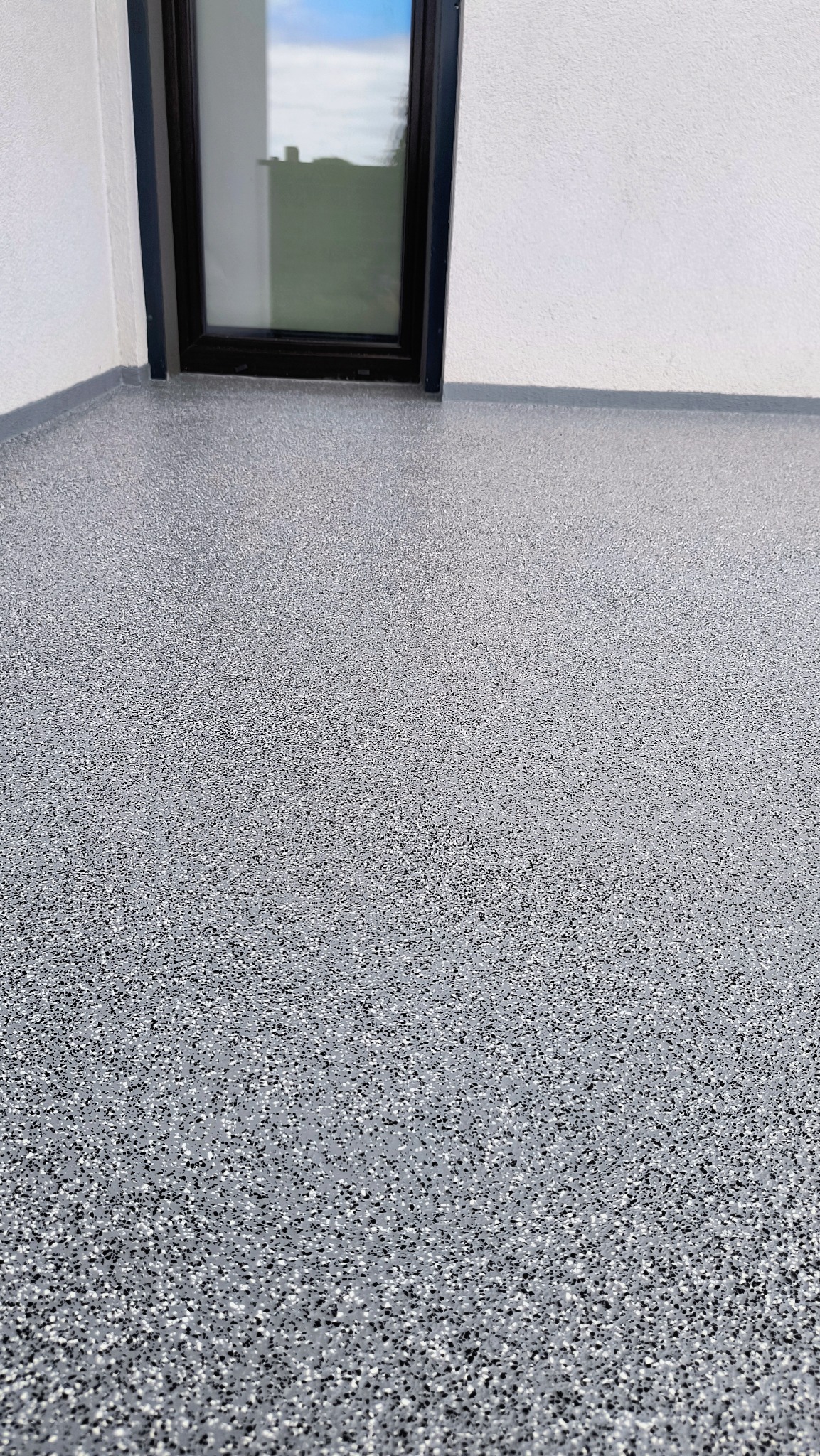 Szara posadzka żywiczna z czarnymi i białymi drobnymi elementami, widok z góry, przy białej ścianie z oknem.