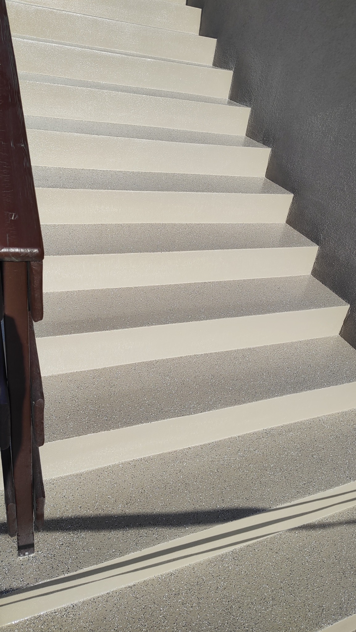 Schody wewnętrzne pokryte beżową, drobnoziarnistą powłoką, z ciemnobrązową metalową barierką po lewej stronie.