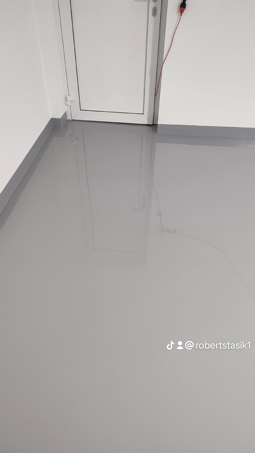 Gładka, szara podłoga z wylewki samopoziomującej w nowym pomieszczeniu, z widocznym progiem i białą ścianą z widocznym kablem elektrycznym.