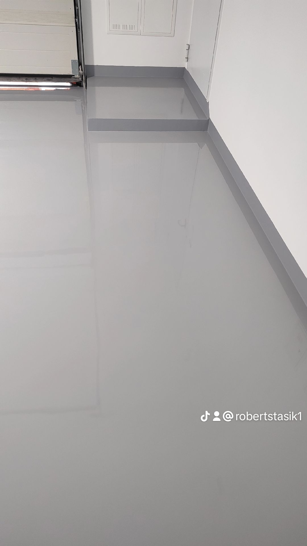 Jasnoszara, błyszcząca posadzka żywiczna w garażu, widoczny fragment bramy garażowej i biała ściana z szarą listwą przypodłogową, ujęcie z góry pod kątem.