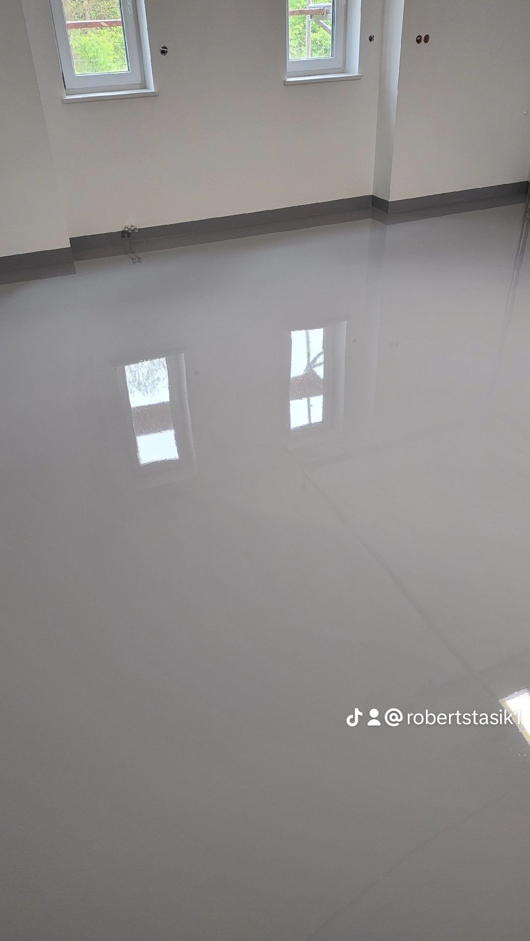 Szara, błyszcząca podłoga w nowym pomieszczeniu z dwoma oknami, odbijającymi się w powierzchni. Widoczne ślady po instalacji elektrycznej na ścianie.